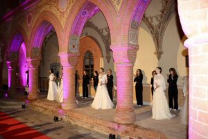 Una sfilata di moda in un antico loggiato del Castello di Cereseto: un lungo tappeto rosso steso lungo un corridoio all'aperto affiancato da arcate in mattoni decorate con affreschi. Un gruppo di modelle indossa eleganti abiti da sposa bianchi di vari stili.